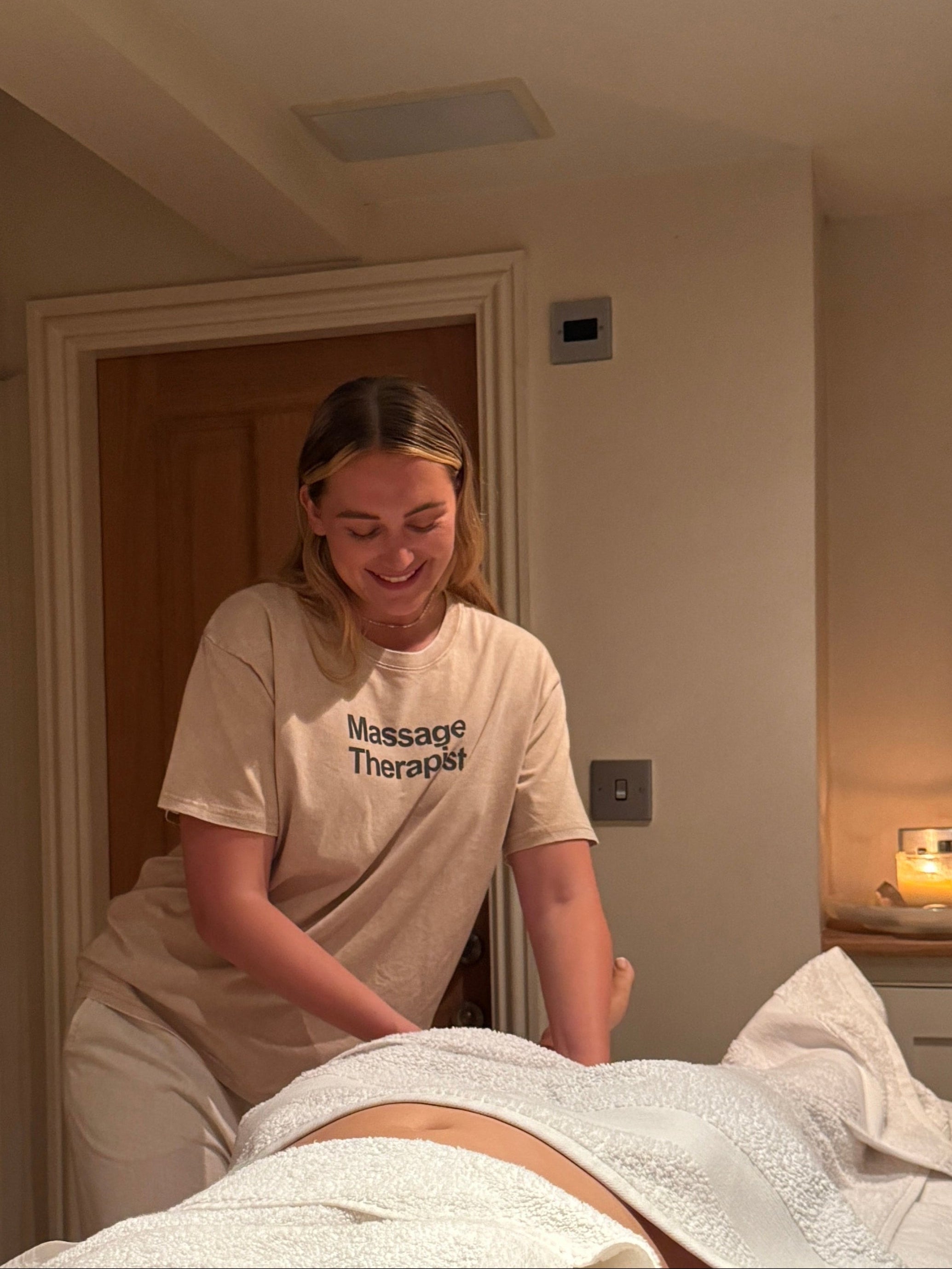Massage therapist performing a  lymphatic drainage massage in a room with a candle and decorative items on a shelf.